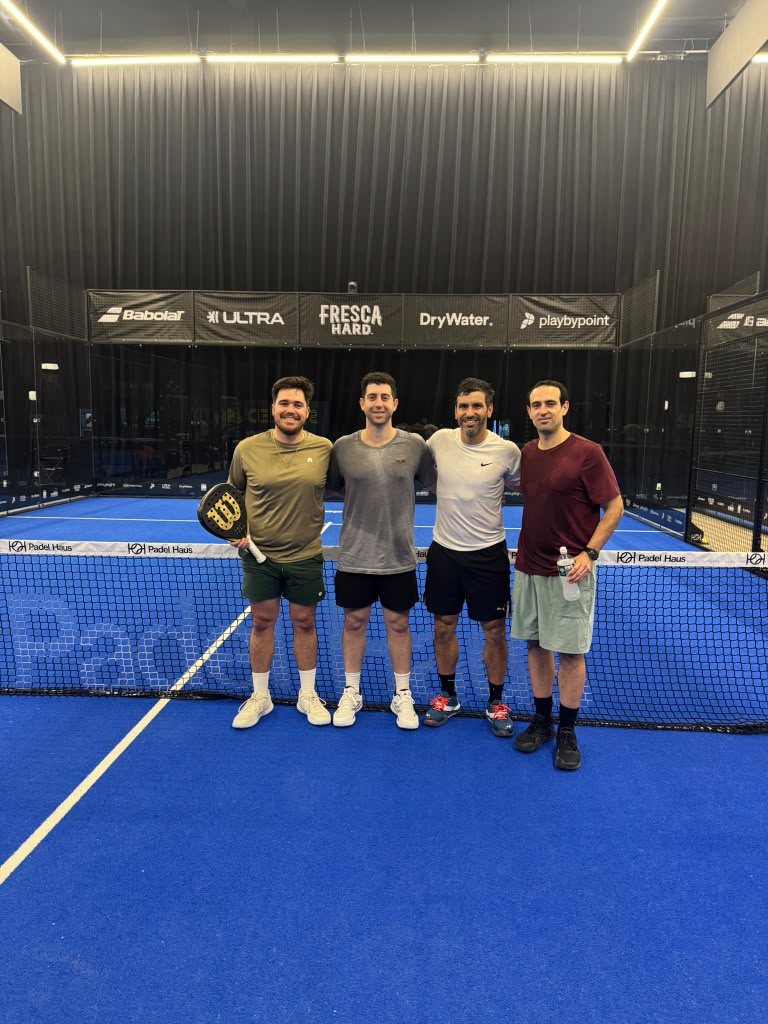Four players posing at the net on a blue indoor court at Padel Haus